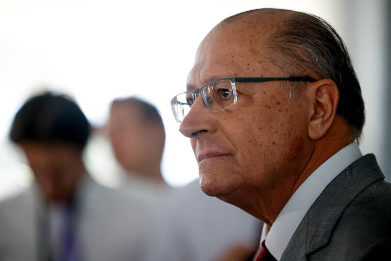 Alckmin, homem branco de óculos, está de perfil esquerdo na metade direita da imagem. Ao fundo, duas pessoas desfocadas