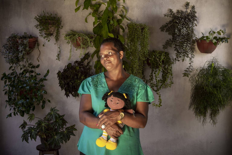 Imagem mostra Madalena Gordiano segurando uma boneca de tricô, que ganhou de um promotor federal logo após ser resgatada de condições análogas à escravidão. Madalena é uma mulher negra e veste uma blusa verde e está em um ambiente com plantas ao fundo.
