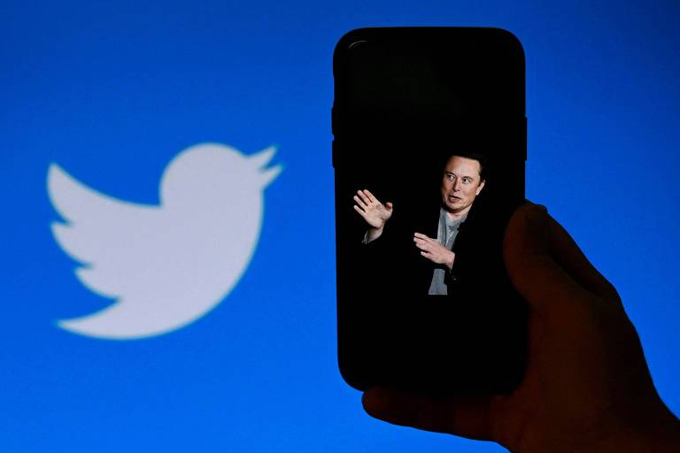 Imagem mostra silhueta de uma mão segurando um celular, cuja tela mostra foto de Elon Musk --um homem branco, de camiseta cinza e jaqueta preta. Ele é branco de cabeços castanhos. Ao fundo, há um banner do Twitter