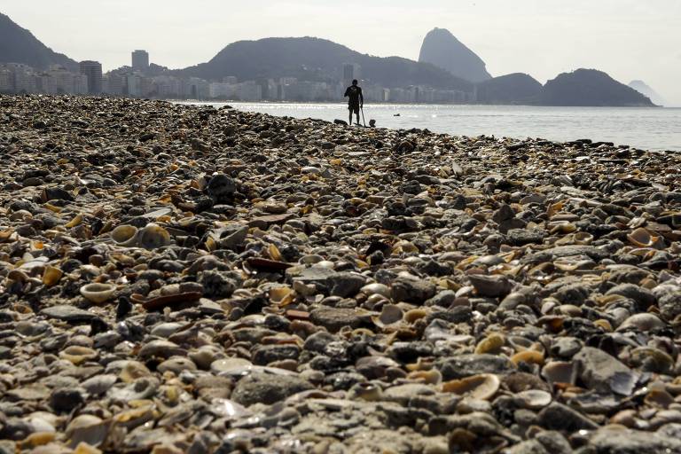 Faixa de areia da praia coberta de pequenas pedras, conchas e sedimentos marítimos. No fundo da imagem, um homem caminha à beira do mar.