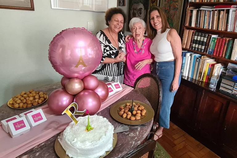 Rosa Cass ao centro, com as sobrinhas Ana Léa à esquerda e Ester à direita. a foto foi tirada no aniversário, há um bolo e decorações diversas com balões 