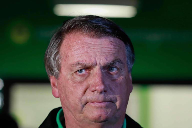 Rosto de Jair Bolsonaro, um homem branco, vestido de preto e verde, mesmas cores que aparecem no fundo da imagem, em desfoque. Ele tem expressão séria e olha para a esquerda
