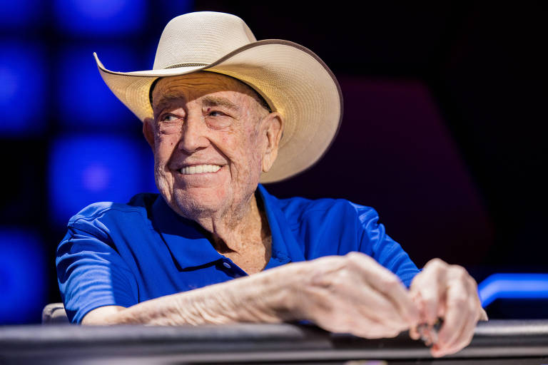 Homem branco de camisa azul e chapéu branco de cowboy sorri com fichas de pôquer nas mãos, sentado numa mesa de jogo