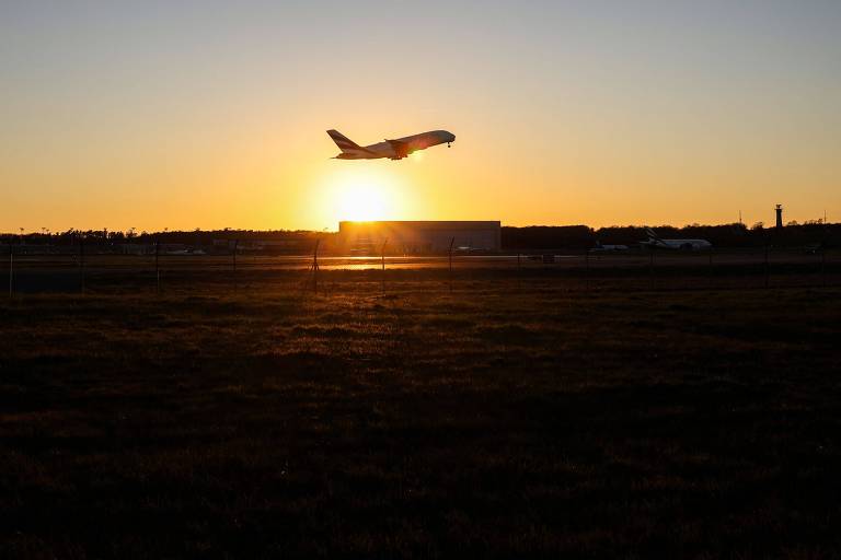 Fotografia colorida mostra, de longe, um avião decolando; atrás dele, é possível ver o pôr do sol