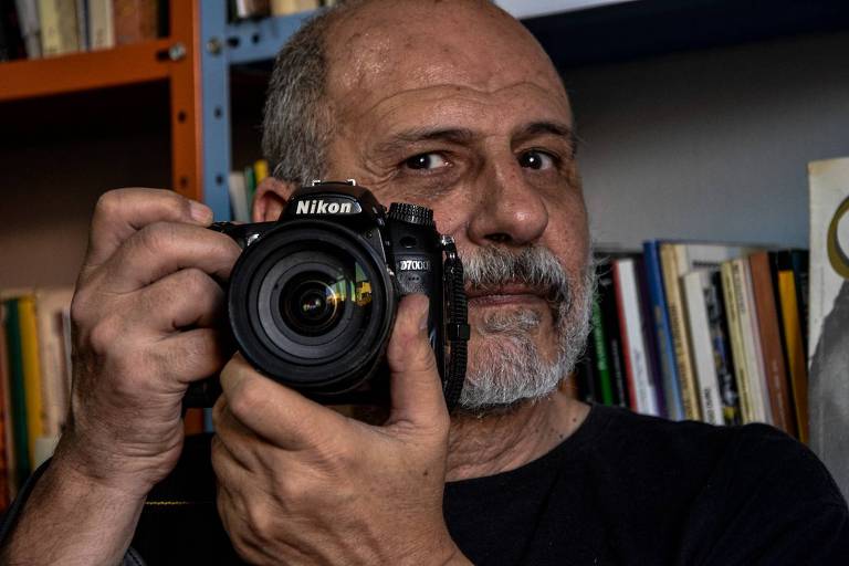 O repórter fotográfico Ennio Frederico Brauns Filho segura uma câmera em frente ao rosto com uma estante de livros ao fundo
