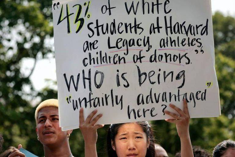 Ao lado de jovem negro, mulher com traços asiáticos segura cartaz com dado de que 43% dos estudantes brancos de Harvard recebem a admissão por "legado" e de outros tipos