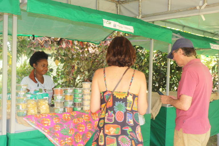 Foto mostra clientes comprando em barraca na Festa da Ostra. A barraca é verde com toalha vermelha na mesa e é comandada por uma mulher negra de blusa branca