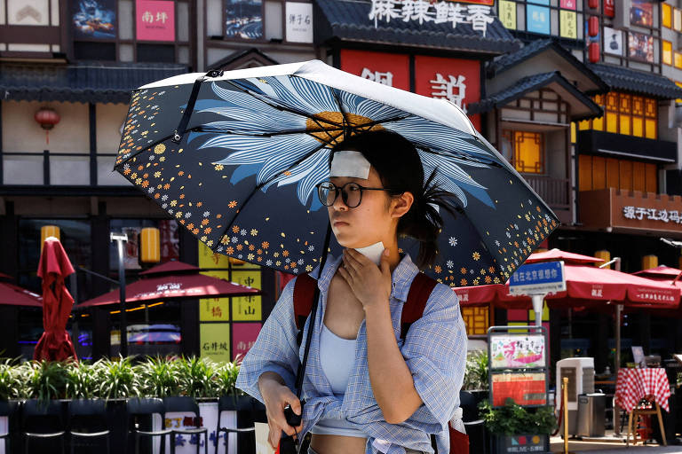 Mulher jovem caminha em uma rua usando uma sombrinha; na pele da testa e do pescoço ela tem coladas bandagens brancas