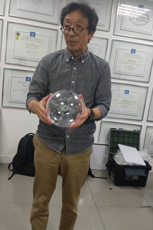 Kikuo Abe, um homem de 71 anos, cabelos pretos e óculos, segura uma esfera com um visor de relógio preso a quatro elásticos. Ele veste camisa cinza e calça caqui. Está à frente de parede com vários prêmios Esso pendurados na parede