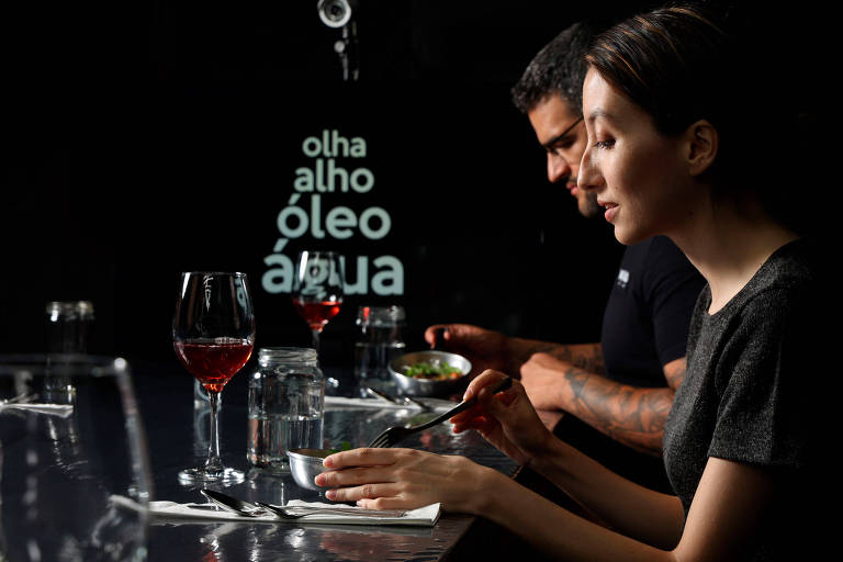 A foto mostra duas pessoas pessoas comendo em um evento de gastronomia e arte, em um cenário escuro