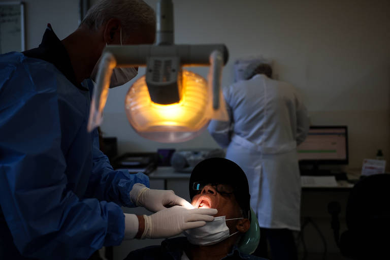 Homem deitado em cadeira de consultório abre a boca enquanto é examinado por dentista