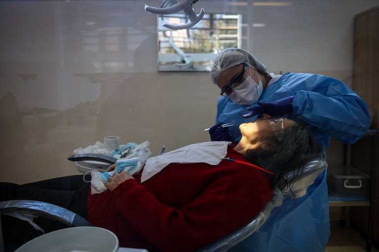 A imagem mostra uma consulta odontológica em andamento. Um dentista, vestido com um avental azul e máscara, está realizando um procedimento em um paciente que está deitado em uma cadeira de dentista. O paciente, uma mulher idosa com cabelo grisalho, usa um suéter vermelho e está com a boca aberta. O ambiente é iluminado e há equipamentos odontológicos visíveis ao fundo.