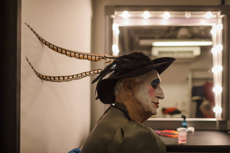 Dramaturgo Zé Celso aparece de perfil, em frente ao espelho em um camarim, com chapéu adornado por duas longas penas