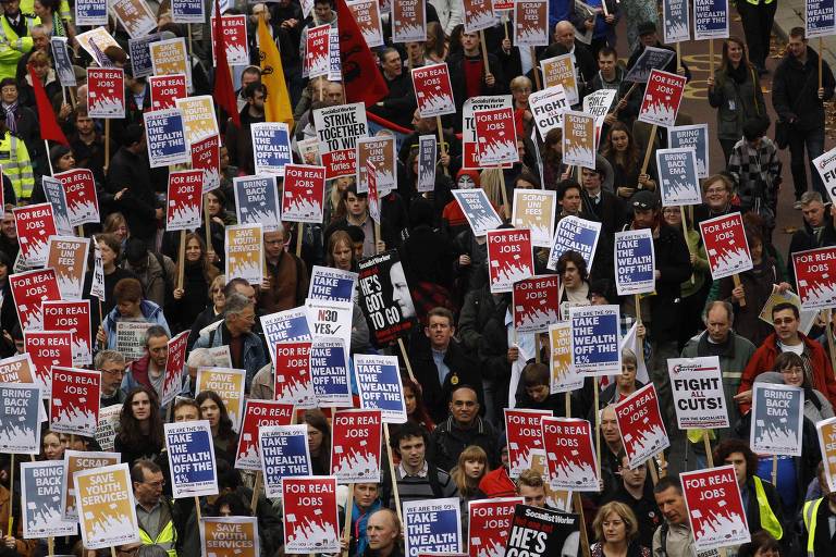Segurando cartazes vermelhos, azuis e amarelos, manifestantes protestam contra corte de empregos na Inglaterra
