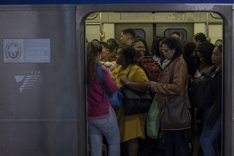 Mulheres em pé dentro de um vagão de trem do Rio de Janeiro. Externamente, o vagão é metálico, de cor prata. Dentro, um grupo de mulheres dividem espaço e aguardam a partida do trem. A porta de embarque do trem está aberta
