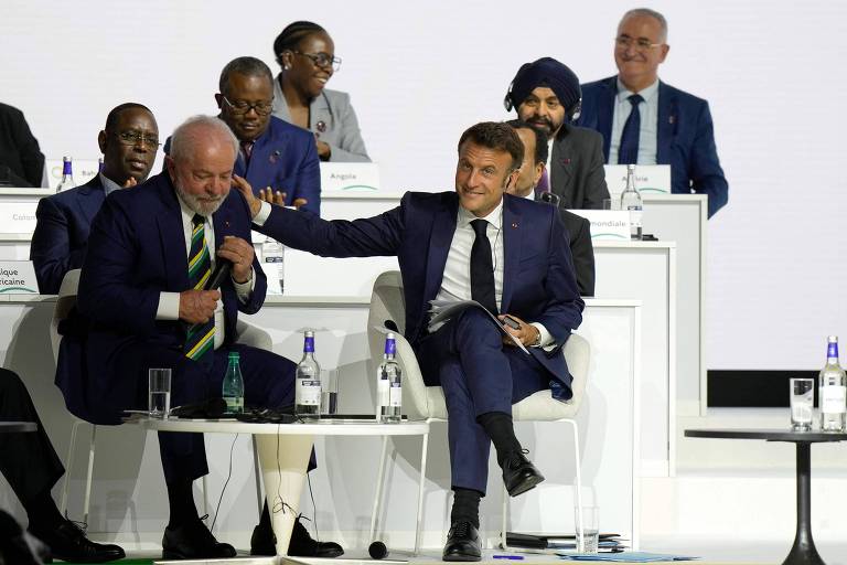 Lula está sentado à esquerda da foto, segurando microfone, enquanto Macron, a sua direita, está com o braço direito esticado com a mão no ombro de Lula. Ao fundo, outras pessoas sentadas. O ambiente é branco.