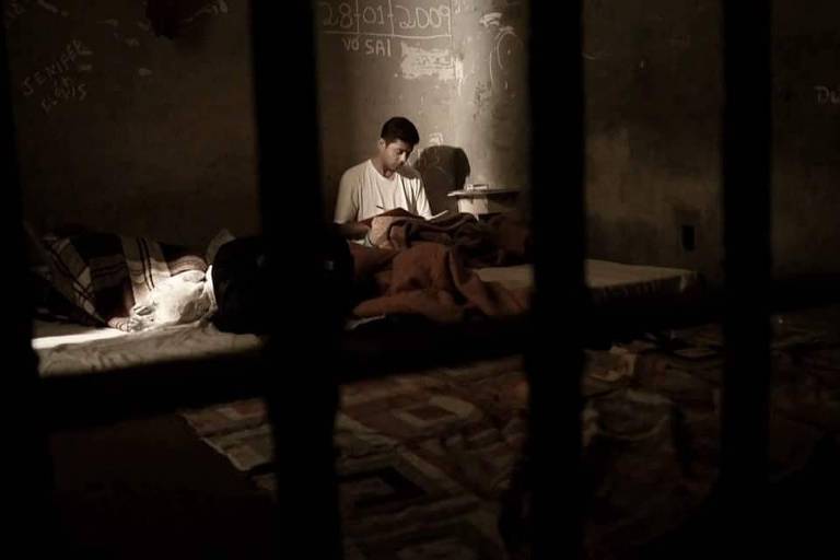 Imagem mostra grades. Ao fundo, o escritor Paulo Milhan está sentado sobre um colchonete posicionado no chão. Ele lê um livro
