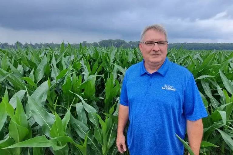 Homem branco vestindo camiseta azul em pé em meio a uma plantação de milho