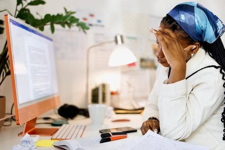 Mulher negra, de longos cabelos cacheados, veste camisa branca e lenço azul, leva a mão à cabeça, em sinal de estresse. À sua frente está um computador, que exibe um editor de texto na tela.