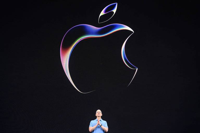 Ceo da Apple aparece em frente a um telão com a logo da empresa, que é uma maçã mordida. Ele está com as mãos unidas.
