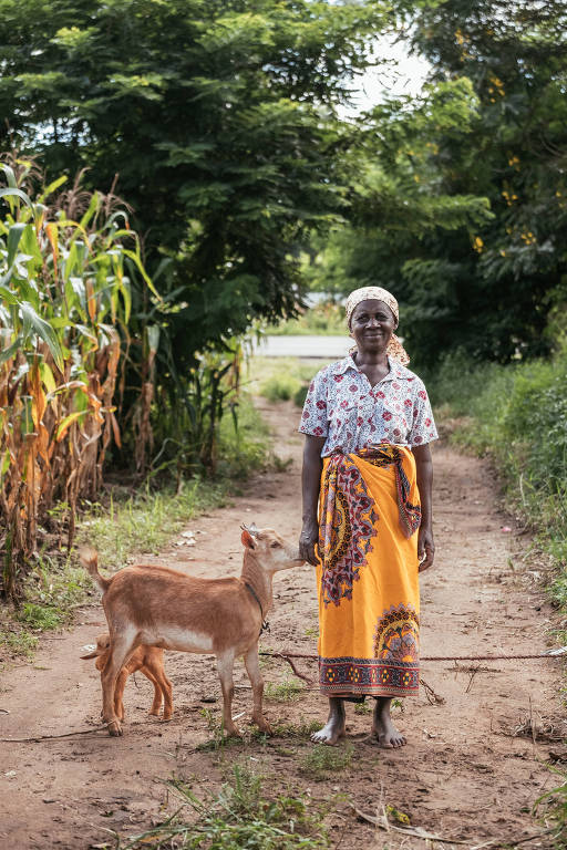 Mulher negra posa ao lado de uma cabra em uma trilha de terra, com árvores ao fundo
