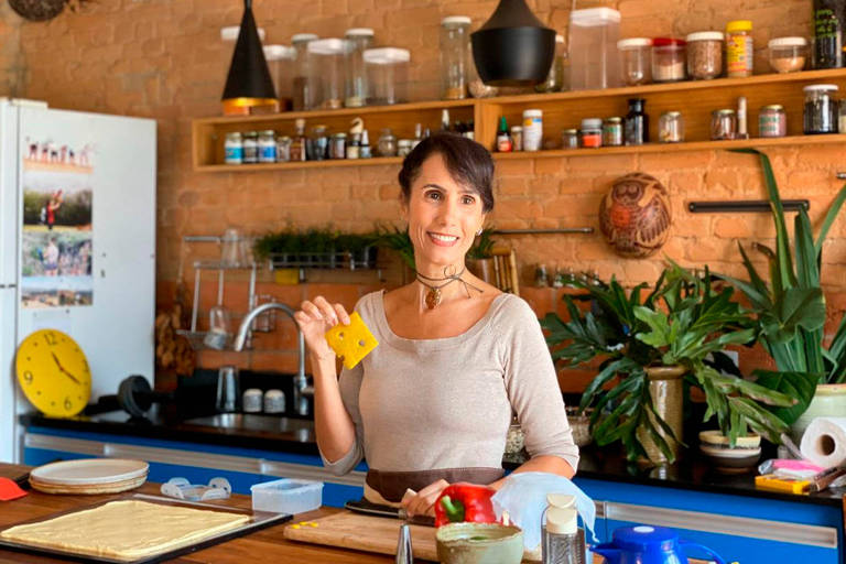A foto mostra uma mulher atrás de uma bancada de cozinha, segurando uma fatia de queijo na mão.