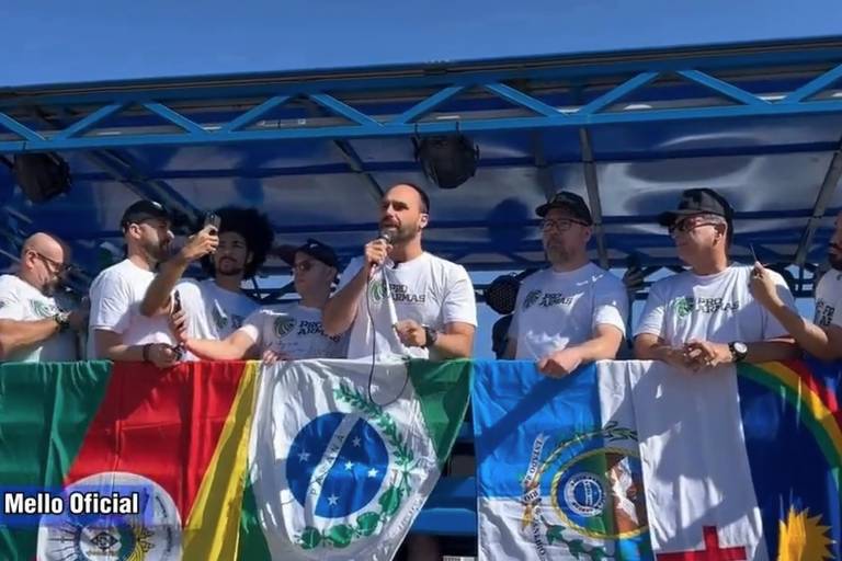 Eduardo Bolsonaro, ao centro, discursa em palco decorado com bandeiras, ao lado de outras pessoas