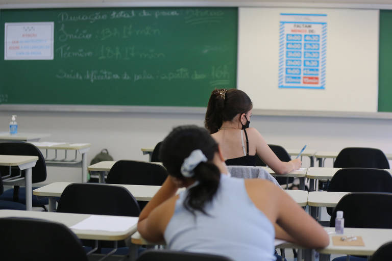 Estudantes sentadas em carteiras e escrevendo em papel são vistas de costas
