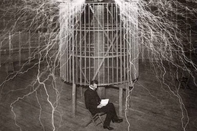 foto mostra tesla sentado lendo um livro à frente do transmissor, é possível ver raios de luz saindo da estrutura