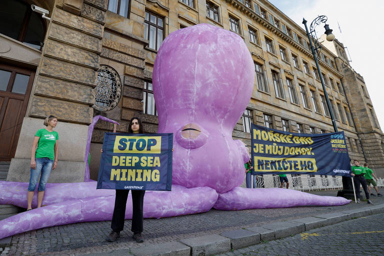 Pessoas seguram cartazes em frente a um edifício com um polvo inflável gigante, cor de rosa, montado em sua calçada