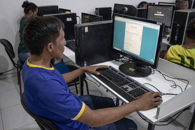 Foto mostra cerca de quatro computadores disponibilizados em quatro estações de trabalho individuais; nos assentos estão três alunos, sendo um homem, uma mulher e um garoto; eles operam os computadores e parecem participar da aula