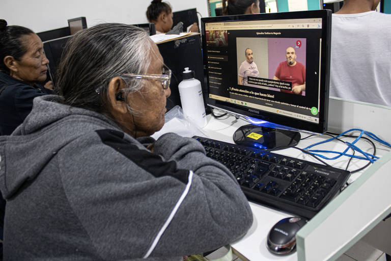 Homens e mulheres venezuelanos estão sentados à frente de computadores individuais e assistem a vídeo; na foto aparecem cerca de cinco alunos