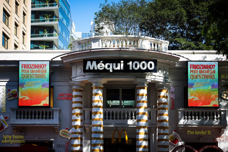 prédio branco onde fica loja da rede McDonald's. No topo, sobre a entrada principal, há uma placa o nome do restaurante: Méqui 1000. Dos dois lados, telões de led convidam visitantes a entrar e ofertam produtos