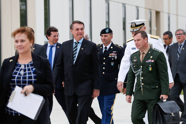 O ex-presidente Jair Bolsonaro, um homem branco de terno azul, e Mauro Cid, um homem branco de cabelo curto e farda verde do Exército, caminham junto a outras pessoas durante viagem nos Estados Unidos.