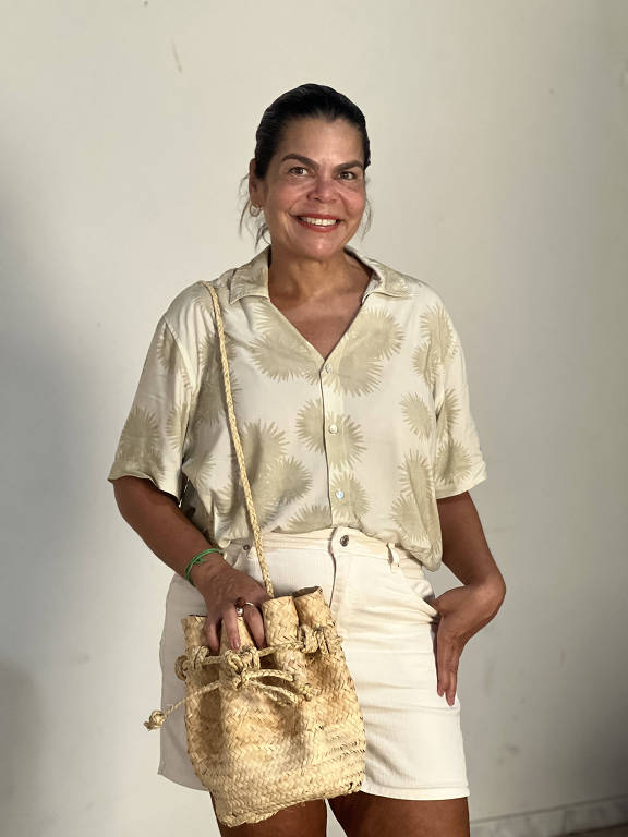 Mulher sorri para a foto mostrando uma bolsa feita de palha de carnaúba; em pé, ela usa uma camisa clara estampada com folhas verdes e um shorts branco