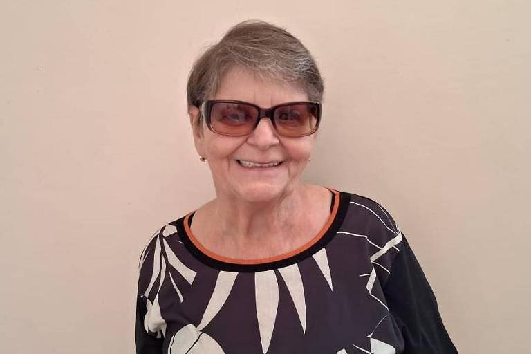Sônia, 67, sorri em frente a uma parede beige; sua pele é branca, ela tem cabelos grisalhos e curtos e usa óculos escuros, uma blusa