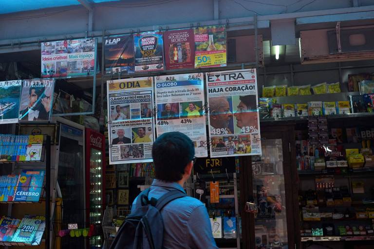 Fotografia colorida em plano médio de homem parado diante de banca de revistas; ele está de costas para o fotógrafo e observa o conteúdo exposto