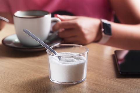 Woman with clock and phone holding a cup of coffee with milk and next to sugar substitute in a cafe.Credit Zakiroff / AdobeStock