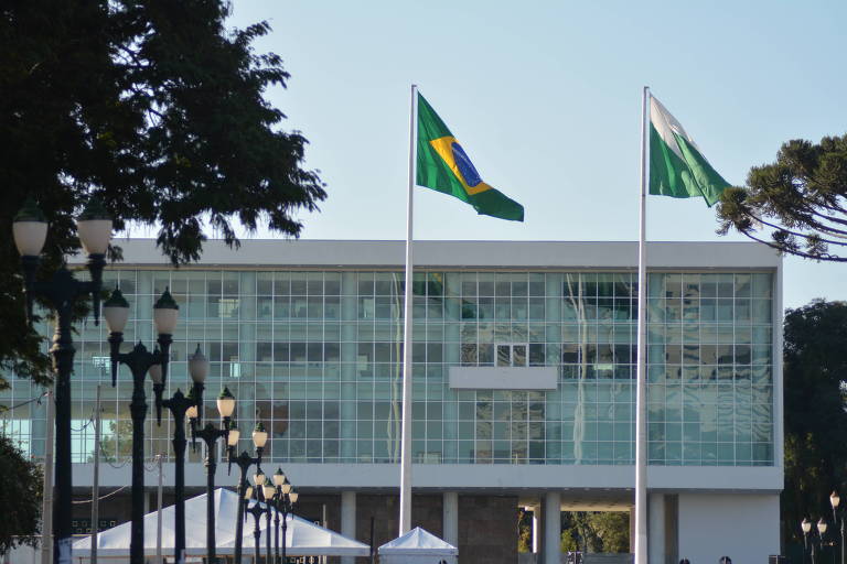 Imagem mostra palácio do Iguaçu, um prédio espelhado de paredes brancas. É possível visualizar árvores e postes de luz no lado esquerdo e, ao centro e no lado direito, as bandeiras do Brasil e do estado do Paraná, respectivamente