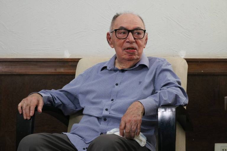 Homem idoso com camisa azul, óculos, sentado em uma poltrona e os braços apoiados nela