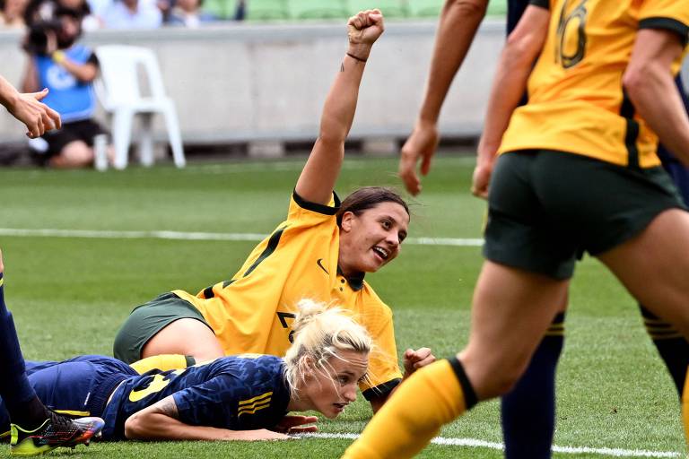 Kerr, de camisa amarela e shorts verde da seleção australiana, está sorrindo e deitada, com o braço direito erguido para cima. Uma jogadora da Suécia está caída ao seu lado 