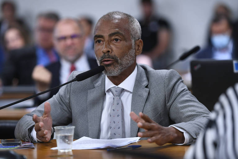 Romário, um homem careca e de barba branca, fala durante audiência pública a outros parlamentares. Ele aparece sentado e veste terno cinza.