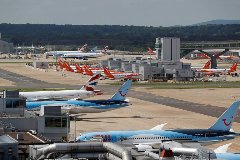 Imagem de um aeroporto com várias aeronaves estacionadas. No primeiro plano， há aviões de uma companhia aérea com a cauda azul e branca. Ao fundo， várias aeronaves de diferentes companhias estão alinhadas na pista， com destaque para as caudas vermelhas e laranjas. O céu está claro e há uma área de terminal visível.