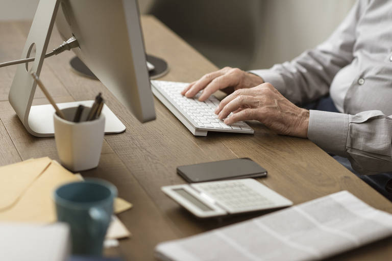 Mãos de homem digitando em teclado sem fio; sobre a mesa há  celular, jornal, caneca, canetas e uma calculadora