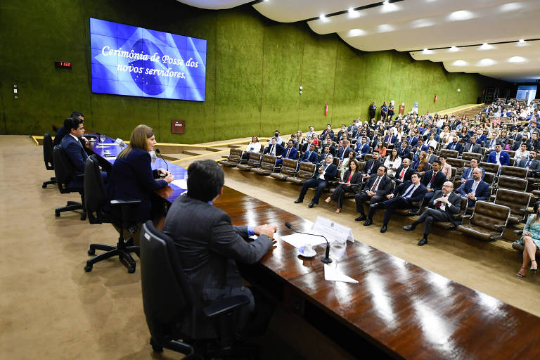 Imagem mostra pessoas em auditório da Câmara dos Deputados; no telão, ao fundo, está escrito: "Cerimônia de Posse dos novos servidores"