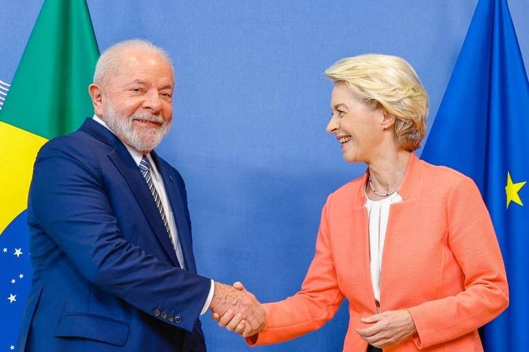   Imagem mostra Lula apertando a mão de Ursula. os dois sorriem. Atrás, estão a bandeira do Brasil e a bandeira da UE