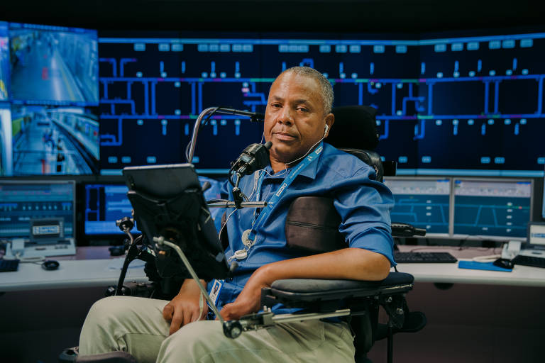Um homem de camisa azul está em um cadeira de rodas motorizada e com comandos na altura de sua boca; atrás dele, um painel com vários monitores