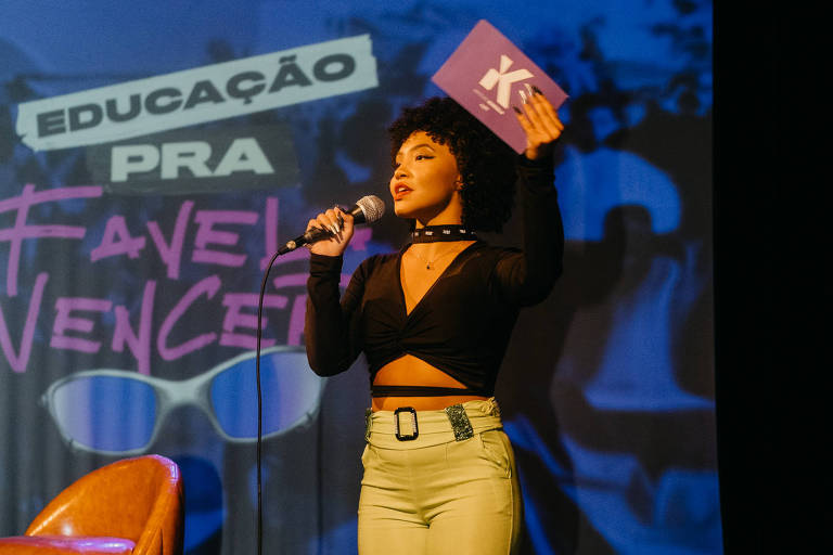 Mulher negra fala ao microfone em um palco durante evento; ela usa blusa de cor preta e calças de cor bege