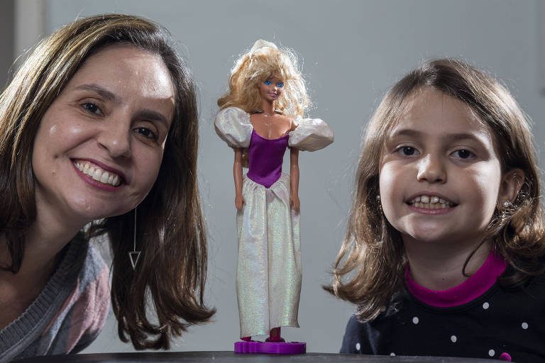 Mãe e filha olham para a câmera; entre elas, em cima da mesa, está uma Barbie loira com vestido de princesa