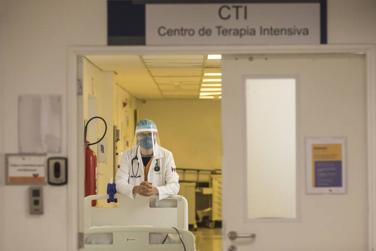 A foto mostra uma fisioterapeuta com máscara e faceshield de frente para a câmera na porta de uma sala de CTI de hospital. Acima da porta há uma placa onde se lê: Centro de Terapia Intensiva